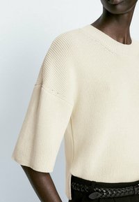 JasnopBeżowy sweter dziany z krótkimi rękawami, o prążkowanej teksturze i luźnym kroju. Ma dekolt w łódkę i boczne rozcięcia.