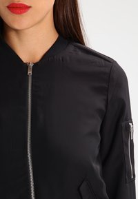 Black bomber jacket with zip closure, ribbed collar, and side pockets. Features a zippered pocket on the sleeve. Smooth, lightweight material.