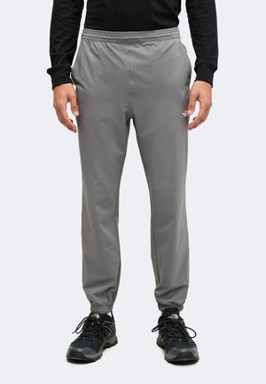 Mann trägt graue Jogginghosen mit elastischem Bund und elastischen Bündchen sowie schwarze Wanderschuhe, steht vor hellem, einfarbigem Hintergrund.