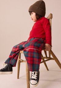 Rotes Langarmshirt, karierte Hose in Rot und Grün und schwarze High-Top-Sneaker. Sitzt auf einem Holzstuhl und trägt eine braune Strickmütze.