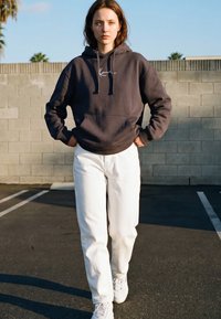 Jeune femme en sweat à capuche noir, pantalon blanc et baskets, debout sur l'asphalte, les mains dans les poches, contre un mur en béton.
