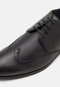 Zapato de cuero negro con una textura lisa y perforada, punta afilada y acentos de brogue detallados a lo largo de los bordes y costuras.