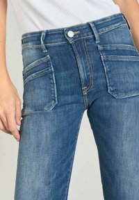 Jean en denim bleu avec poches avant visibles, fermeture à bouton et coutures orange. Dispose d'une coupe droite et d'une texture décolorée.