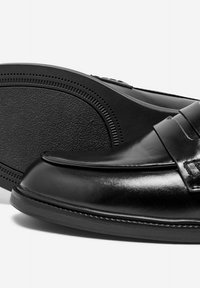 Mocasines de piel negra con un acabado suave, que presentan una correa decorativa y una suela de goma negra texturizada. Detalles de costura mínimos.