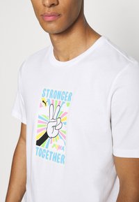 Bílý tričko s krátkým rukávem, které má barevný grafický potisk ruky dělající znamení míru se textem "SILNĚJŠÍ SPOLU" a logem Puma.