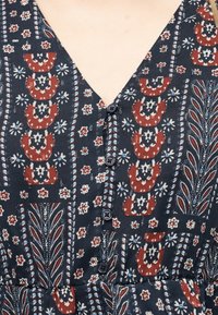 Νavy μπλε ρούχο με V λαιμόκοψη σε κόκκινες, λευκές και ανοιχτό μπλε λουλουδένιες και γεωμετρικές σχεδιάσεις, με μια σειρά από μαύρα κουμπιά κατά μήκος του μπροστινού μέρους.