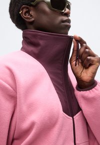 Άτομο που φοράει μεγάλα γυαλιά ηλίου με πράσινες αποχρώσεις και ένα ροζ μπουφάν με ψηλό σκούρο μωβ γιακά, το οποίο κλείνει με φερμουάρ με το χέρι του.