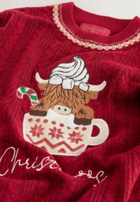 Sarkans adīts džemperis ar izšūtu brūnu govs attēlu, kas sēž baltā krūzē ar krēmkrāsas virsotni, papildināts ar saldumu nūjiņu un sniegpārsliņu rakstiem.