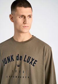 Camiseta de algodón verde oliva con el texto grande en azul marino "JUNK de LUXE" y un gráfico de una corona, escote redondo y corte relajado.