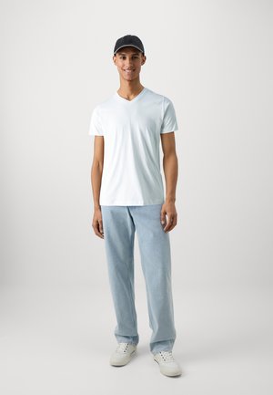 Hvit kortermet V-hals t-skjorte, lyseblå jeans og hvite joggesko. Modellen har på seg en mørk caps og står mot en ensfarget bakgrunn.