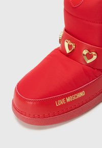 Red padded boot featuring a smooth surface, gold heart-shaped accents, and embossed logo detail along the side. Flat rubber sole.
