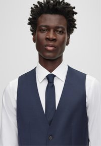 Marineblaue Weste mit glatter Textur über einem weißen Hemd, kombiniert mit einer passenden Krawatte. Verfügt über einen einzelnen Knopf und schräge Taschen.