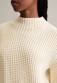 Helle beige Strickjacke mit hohem Kragen, die mit einem strukturierten Muster aus kleinen, erhabenen Punkten und gerippten Details versehen ist.