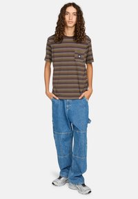 T-shirt rayé marron, jaune et violet avec une poche sur la poitrine ; associé à un jean en denim léger, bleu clair et ample avec un design en patchwork.