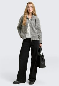 Vrouw met lang blond haar, gekleed in een grijze ritsjack, een witte shirt, zwarte wijde broek, zwarte laarzen en een zwarte handtas.