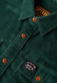 Camicia verde in velluto a coste con colletto abbottonato, caratterizzata da bottoni arancioni e un'etichetta con la scritta "Superdry Co. Machined." Tessuto texturizzato.