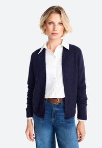 Cardigan blu navy, in tessuto strutturato, con design aperto e polsini a coste, indossato sopra una camicia bianca con bottoni e jeans blue classici.