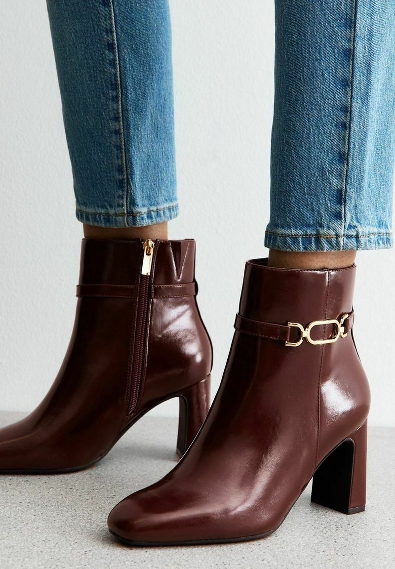 Bottines en cuir brun foncé brillant avec talons carrés, fermeture éclair dorée sur le côté et détail chaîne dorée portées avec un jean bleu.