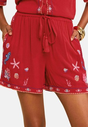 Rote Shorts mit Kordelzug an der Taille und bestickten meeres-themed Designs wie Seesterne, Quallen und Muscheln, getragen mit goldenen Armbändern.