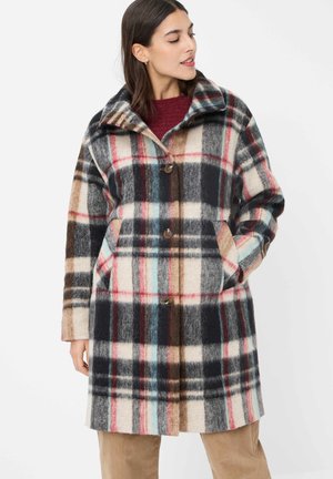 Frau trägt einen knielangen karierten Mantel mit großen Knöpfen und Seitentaschen, darüber einen burgunderroten Pullover und beige Hosen.