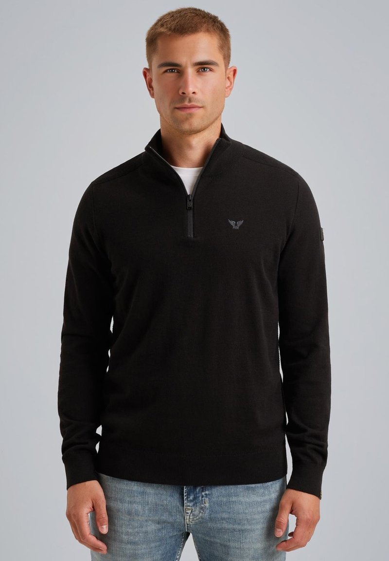 Pull à col zippé noir en matière tricot douce, avec des accents côtelés sur les épaules et un petit logo sur la poitrine. Porté avec un jean bleu clair.