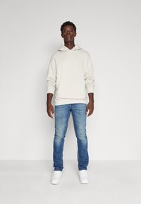 Beiger Hoodie mit Kordelzug und Kängurutasche, kombiniert mit hellblauen Jeans und weißen Sneakern, vor einem einfarbigen Hintergrund stehend.