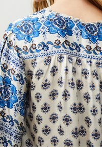 Blusa fluida in tessuto leggero con un design floreale blu su uno sfondo crema, con motivi intricati e dettagli arricciati al collo.