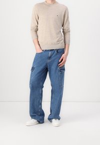 Beige neulepusero, jossa on pyöreä kaula-aukko, yhdistettynä vaaleansiniin cargo-farkkuihin, joissa on sivutaskut ja rento istuvuus, täydentävät valkoiset tennarit.