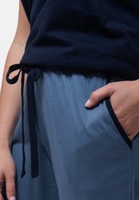 Person som bär blå dragsko-byxor med handen i fickan och en mörkblå topp knuten i midjan.