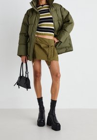 Chaqueta de plumón verde oliva, suéter recortado a rayas negras y oliva, falda oliva con detalles de herrajes, bolso negro y botas de tacón grueso.