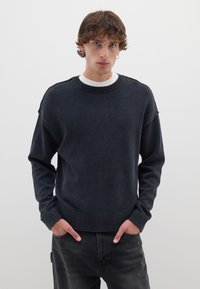 Pullover blu scuro con finitura testurizzata, polsini e orlo a costine; indossato sopra una maglietta bianca a girocollo. Il modello posiziona le mani nelle tasche.