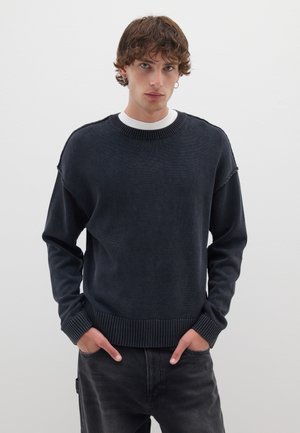 Dunkelblauer Pullover mit strukturiertem Finish, gerippten Bündchen und Saum; getragen über einem weißen Rundhalsausschnitt-Shirt. Das Modell posiert mit den Händen in den Taschen.