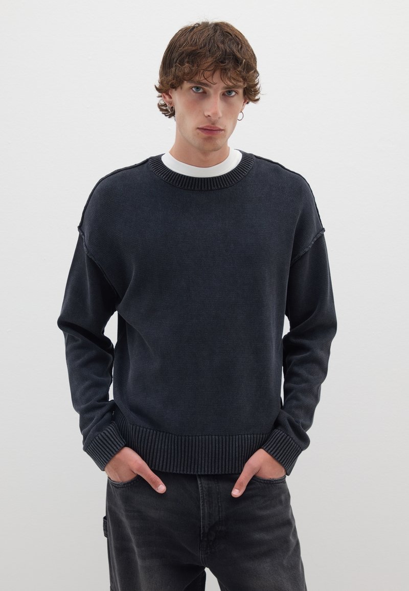 Pullover blu scuro con finitura testurizzata, polsini e orlo a costine; indossato sopra una maglietta bianca a girocollo. Il modello posiziona le mani nelle tasche.