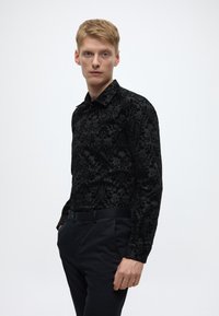 Mustast tekstuurse damaskmustriga pikkade varrukatega särk, millel on terav krae ja kitsas lõige, kombineeritud tumedate pükstega.