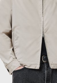 Beige lichtgewicht jack met een frontrits, zijzakken en knopenmanchetten, gecombineerd met donker denim jeans. Vlotte textuur en minimalistisch ontwerp.