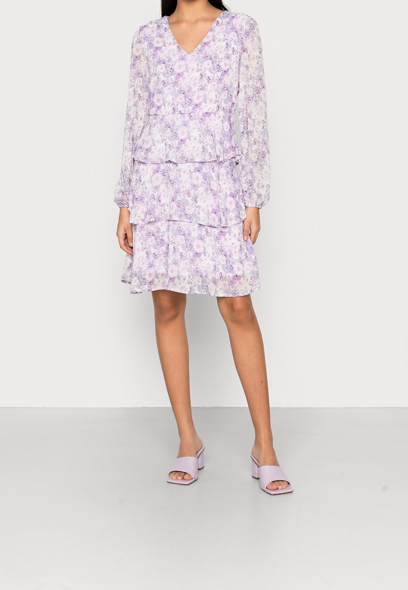 Femme portant une robe florale lavande à manches longues avec décolleté en V, jupe à volants et sandales à talon bloc ouvertes assorties, debout sur un sol gris.