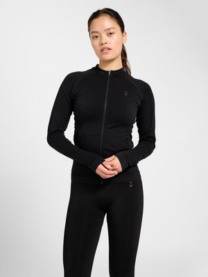 Junge Frau steht vor weißem Hintergrund, trägt eng anliegende schwarze Reißverschlussjacke und schwarze Leggings, die Hände vor der Taille verschränkt.