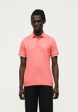 Uomo dalla pelle scura con dreadlocks che indossa una polo rosa corallo con rifiniture bianche e pantaloni neri, in piedi davanti a uno sfondo bianco.