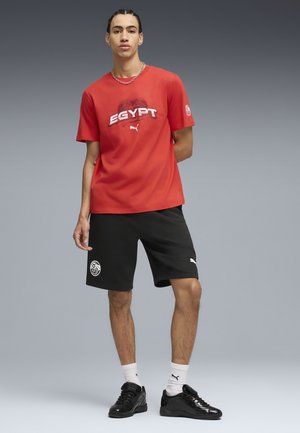 Male model wearing red Puma Egypt t-shirt, black shorts, white Puma socks, and black shoes, standing with a neutral gray background.