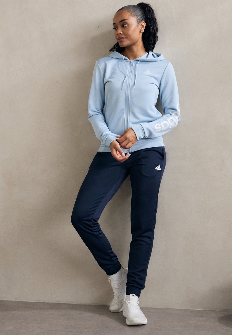 Svetlo modrá mikina na zips s bielou prúžkou a logom na rukáve. Námornícke športové nohavice s priliehavým strihom. Biele športové topánky.