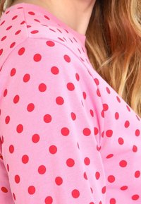 Vaaleanpunainen pitkähiha, jossa on punaisia pisteitä. Kangas näyttää pehmeältä ja sileältä, ja siinä on pyöreä pääntie.