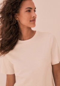 T-shirt color crema con scollo rotondo e maniche corte, realizzata in tessuto morbido, con una texture liscia e un design minimalista.