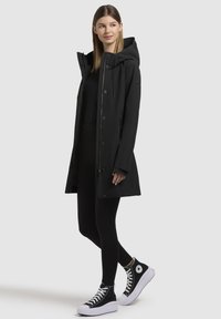 Manteau noir imperméable avec capuche, fermeture à boutons et cordons de serrage ; associé à des leggings noirs et des baskets montantes noires.
