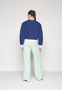 Casaco bomber azul-marinho com mangas bufantes e punhos brancos de ribana, combinado com calças largas verde-menta. Logotipo exibido nas costas.