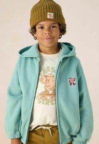 Veste à capuche zippée vert menthe, ornée d'un personnage brodé, portée par-dessus un t-shirt beige clair avec un imprimé de dessin animé et un pantalon marron.