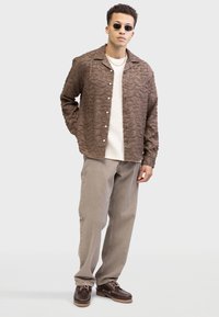 Braun strukturiertes Hemd mit Wellenmuster, cremiger Pullover, beiger Hose und dunkelbraunen Schuhen mit einer kontrastierenden hellen Sohle. Runde Sonnenbrille getragen.