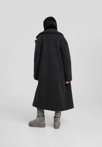 Abrigo teddy negro largo con una tela texturizada y esponjosa, y cuello ancho. Lleva puestos pantalones grises con rayas y botas gris claro.