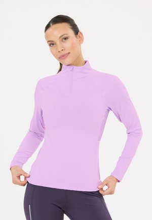 Kvinde justerer kanten på en langærmet lilla kvart-lynlås sportsbluse, iført mørke tætsiddende leggings, foran en ensfarvet hvid baggrund.