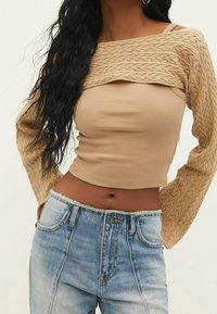 Beżowy prążkowany crop top z teksturowanym, dzianinowym overlayem oraz długimi, wzorzystymi rękawami, zestawiony z jasnoniebieskimi, wysokim stanem dżinsami.