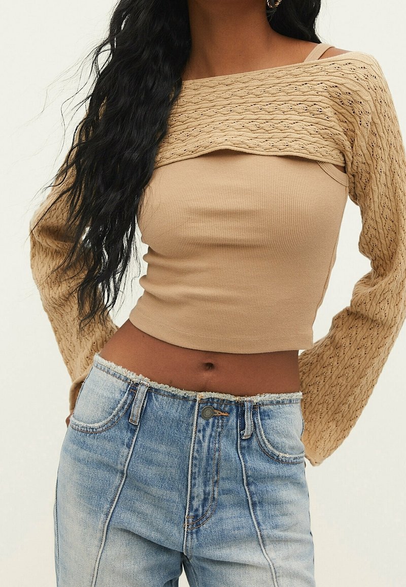 Beżowy prążkowany crop top z teksturowanym, dzianinowym overlayem oraz długimi, wzorzystymi rękawami, zestawiony z jasnoniebieskimi, wysokim stanem dżinsami.